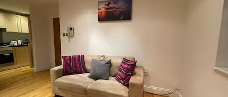 A living room with a beige sofa, two pillows, a painting on the wall, and a kitchen in the background.