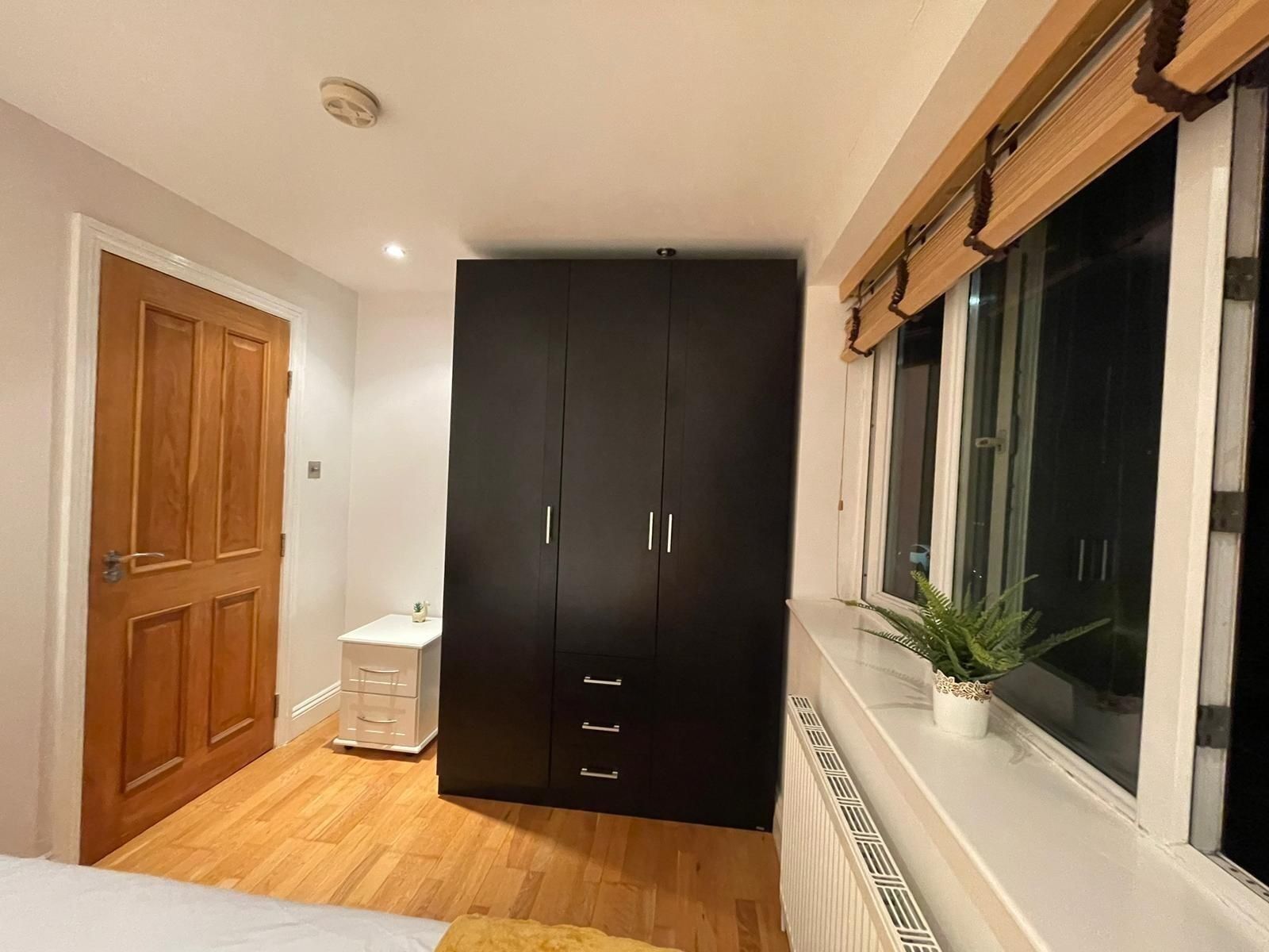 A bedroom with a wardrobe, a bedside table, a window with a plant, and a radiator.