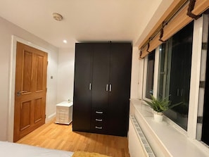 Ein Schlafzimmer mit Kleiderschrank, Nachttisch, Fenster mit Pflanze und Heizkörper.