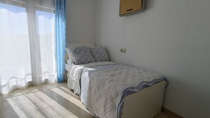 Un llit individual amb llençol estampat, una finestra amb cortines translúcides i un interruptor de llum fixat a la paret.