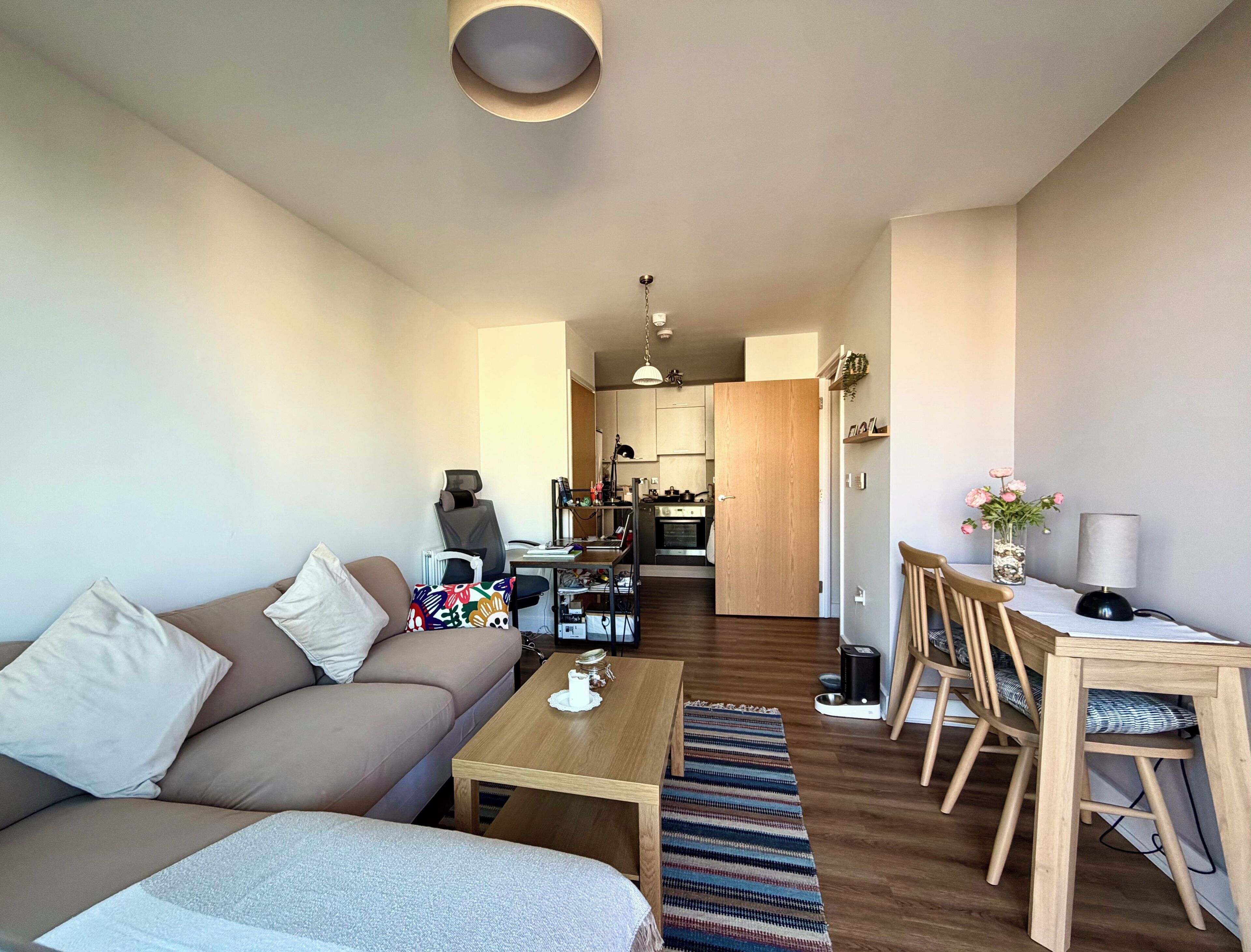Ein modernes Wohnzimmer mit einer Couch, einem Couchtisch und einem Essbereich mit Stühlen und Tisch. Im Hintergrund ist eine Küche zu sehen.