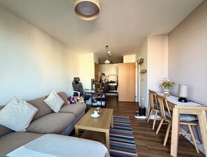 Ein modernes Wohnzimmer mit einer Couch, einem Couchtisch und einem Essbereich mit Stühlen und Tisch. Im Hintergrund ist eine Küche zu sehen.