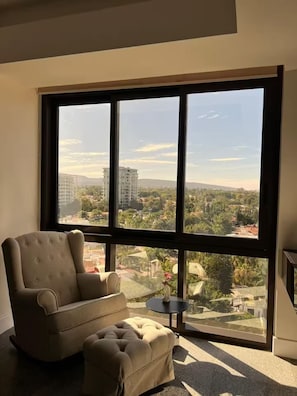 A living room with a large window, a sofa, a small table, and a view of a cityscape.
