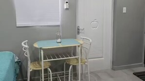 Ein Zimmer mit weißer Tür, einem kleinen Tisch mit Stühlen, einem Fenster mit Jalousien und einem an der Wand befestigten Thermostat.
