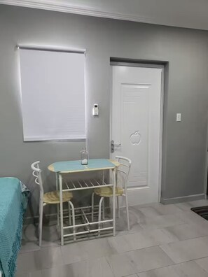 Ein Zimmer mit weißer Tür, einem kleinen Tisch mit Stühlen, einem Fenster mit Jalousien und einem an der Wand befestigten Thermostat.