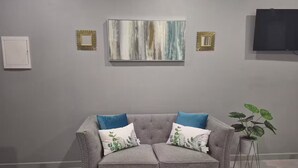 Ein graues Sofa mit zwei dekorativen Kissen, einem an der Wand hängenden abstrakten Gemälde, einem Fernseher und einer Topfpflanze.