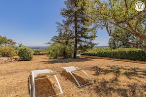 A garden with two white lounge chairs, a large tree, and a view of the countryside.