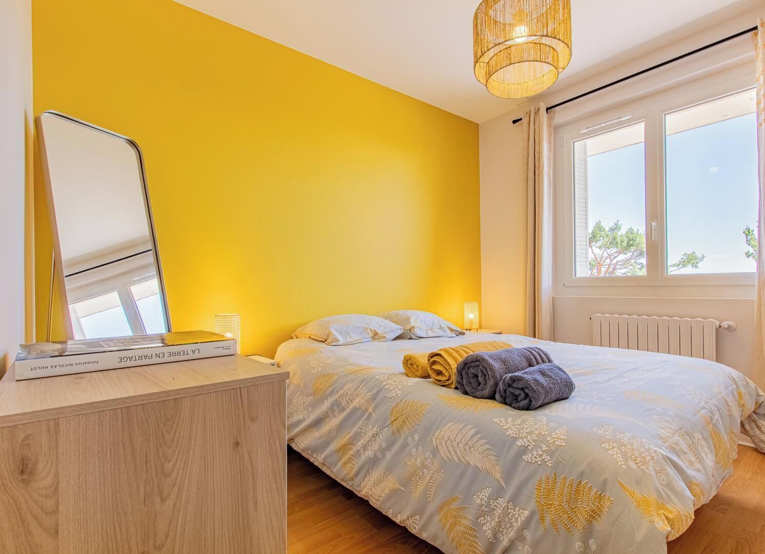 A bedroom with a bed, a bedside table, a mirror, and a window.