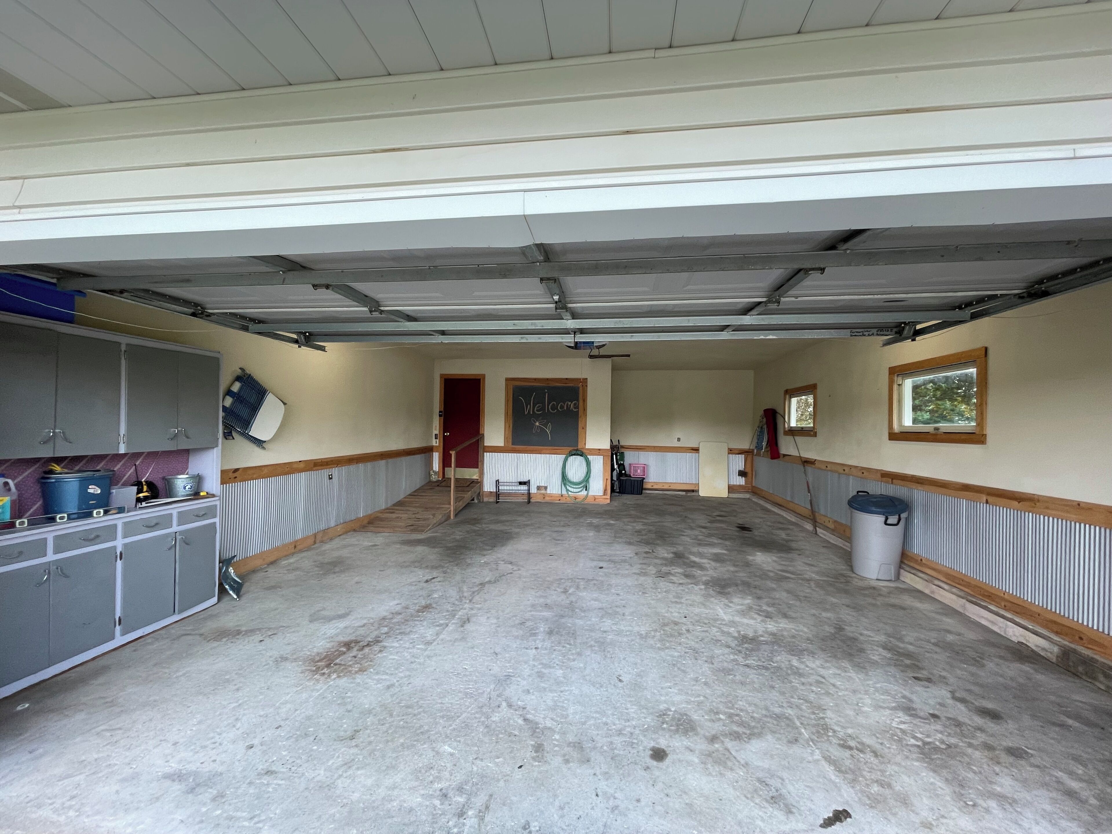 Eine geräumige Garage mit Betonboden, grauen Schränken, einer roten Tür und einem Fenster mit Blick auf Bäume.