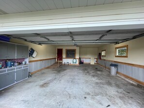 Eine geräumige Garage mit Betonboden, grauen Schränken, einer roten Tür und einem Fenster mit Blick auf Bäume.