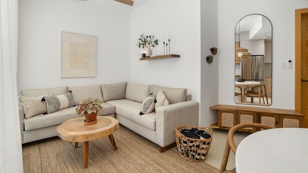Un salon moderne comprenant un canapé, une table basse ronde en bois, un ventilateur de plafond suspendu, une étagère murale agrémentée d’objets décoratifs et un grand miroir qui reflète la pièce.