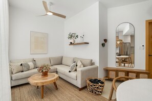 Ein modernes Wohnzimmer mit Sofa, rundem Holztisch, Deckenventilator, einer Wandregal mit Dekoration und einem großen Spiegel, der den Raum reflektiert.