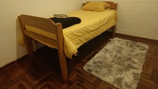 A small room with a wooden bed, a yellow blanket, and a grey rug on a herringbone floor.