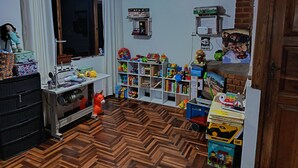A room with a herringbone floor, a sewing machine, a window with curtains, and a bookshelf filled with toys and books.