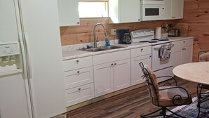 Ett kompakt kök med vita skåp, en diskho, en spis och ett matbord med en stol.