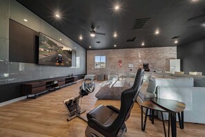 Uma sala de estar moderna com uma televisão de tela plana grande, um simulador de corrida, um sofá cinza e uma parede de tijolos.