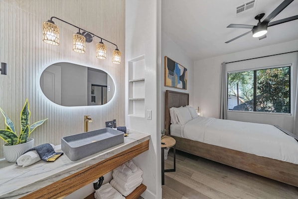 Uma casa de banho moderna com lavatório, pia e espelho. Tem uma cama com vista para o exterior.