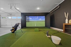 Quarto com grama sintética, uma bolsa de golfe, um balde e uma tela grande exibindo uma vista de campo de golfe.