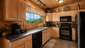 A kitchen with wooden cabinets, a stainless steel refrigerator, a microwave, a coffee maker, and a window with a view of greenery. - Honeymoon VIEW! Beautiful romantic cabin, great location & hot tub w/epic view! (Sevierville)
