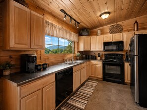 A kitchen with wooden cabinets, a stainless steel refrigerator, a microwave, a coffee maker, and a window with a view of greenery. - Honeymoon VIEW! Beautiful romantic cabin, great location & hot tub w/epic view! (Sevierville)