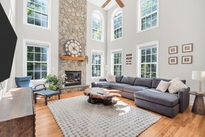 A spacious living room with a stone fireplace, large windows, a grey sectional sofa, and a wooden coffee table. - Private Oasis Retreat with a Pool (Fairfax)