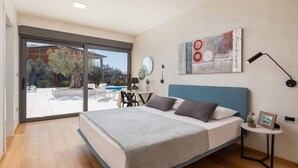 Ein Schlafzimmer mit einem großen Bett, einem Schreibtisch, einem Stuhl, einem kleinen Tisch und Blick auf ein Außen-Schwimmbecken.
