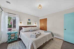 A bedroom with a bed, bedside lamps, a window with curtains, a wooden door, and a small table with a lamp. - New Listing! King Beds, Great Location, Sleeps 6 (Muskegon)