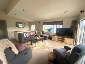 A living room with a sofa, a coffee table, a TV, and a large window. - 53 Snowmass Dr - Ohakune Retreat w/ Ruapehu View (Ohakune)