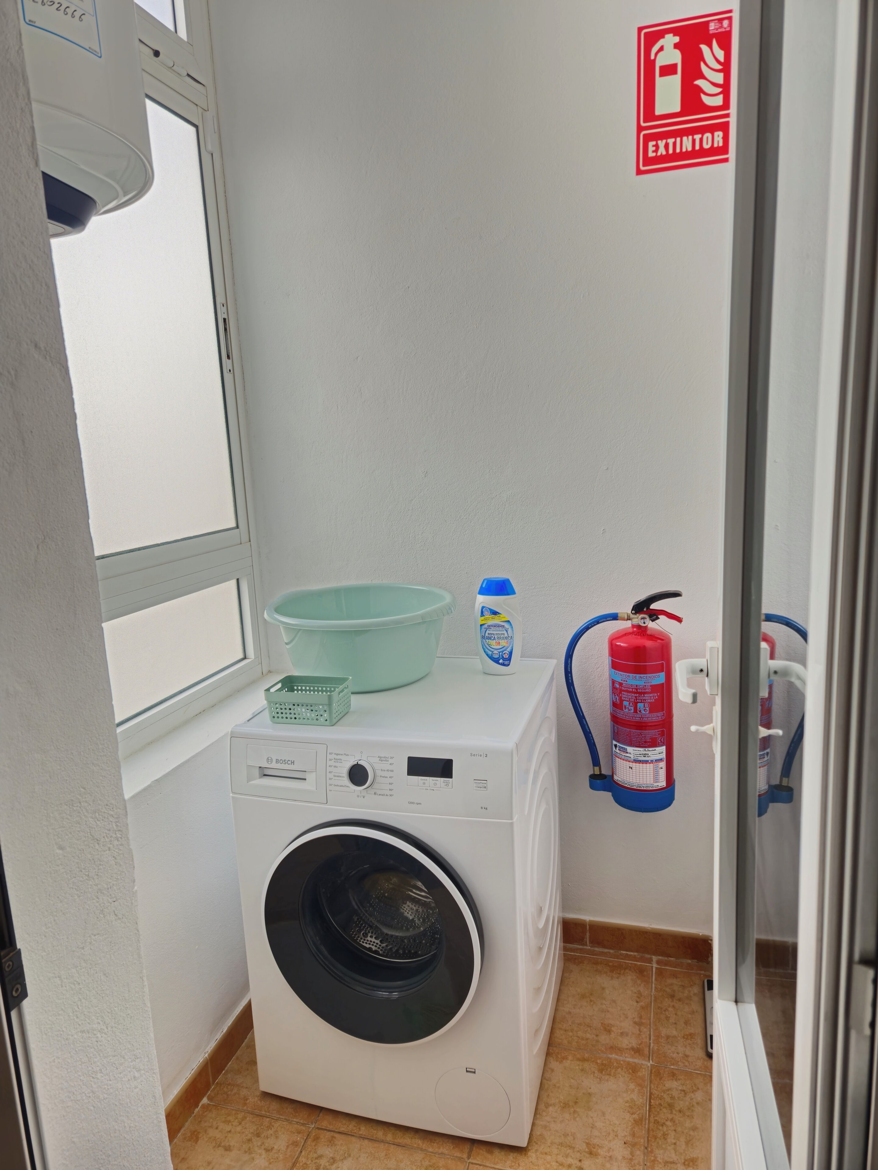 Ein kleiner Waschbereich mit einer Waschmaschine von Bosch, einem Feuerlöscher und einem Fenster.