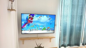 Ett vardagsrum med en platt-TV monterad på väggen, ett litet bord med en dekorativ prydnad, en krukväxt och en gardin.