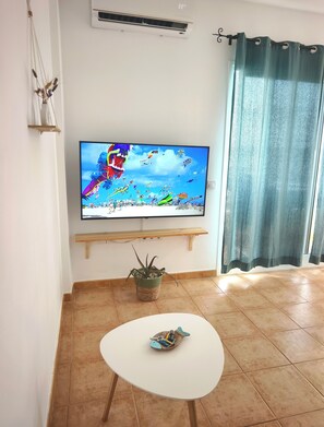 Ein Wohnzimmer mit einem Flachbildfernseher an einer Wandhalterung, einem kleinen Tisch mit einem dekorativen Gegenstand, einer Topfpflanze und einem Vorhang.