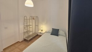 Ett sovrum med en säng, ett sängbord med en lampa, en hylla i metall och en hängande taklampa.