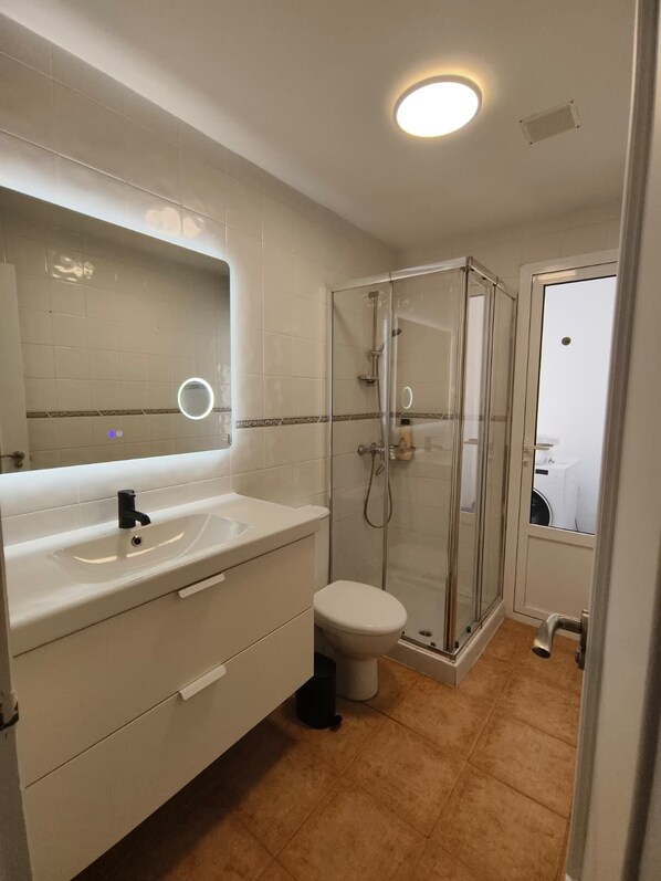 Une salle de bain avec un lavabo blanc, un miroir et des toilettes, une douche en verre et un sol carrelé.