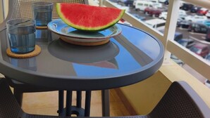En balkong med ett bord, två glas, en tallrik med en bit vattenmelon och utsikt över en parkeringsplats och några byggnader.