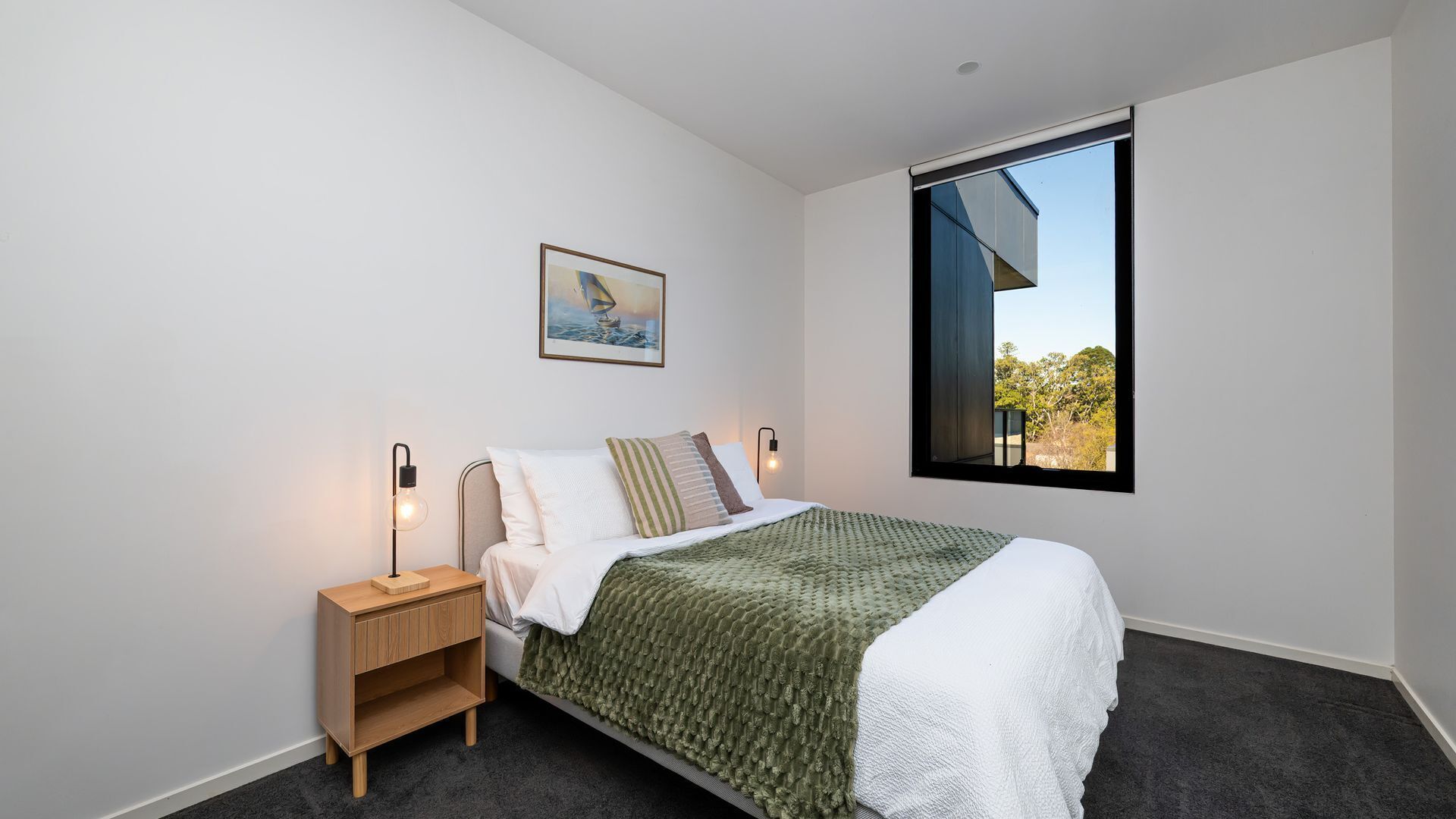Une chambre à coucher comprenant un lit, une table de chevet, une lampe fixée au mur, un tableau et une fenêtre donnant sur un paysage.