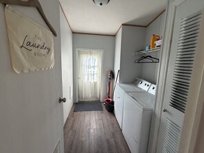 Una sala de bugaderia amb una porta amb la inscripció 'The Laundry Room', una rentadora, una màquina d'assecar i accessoris de neteja com una escombra i un palmell.