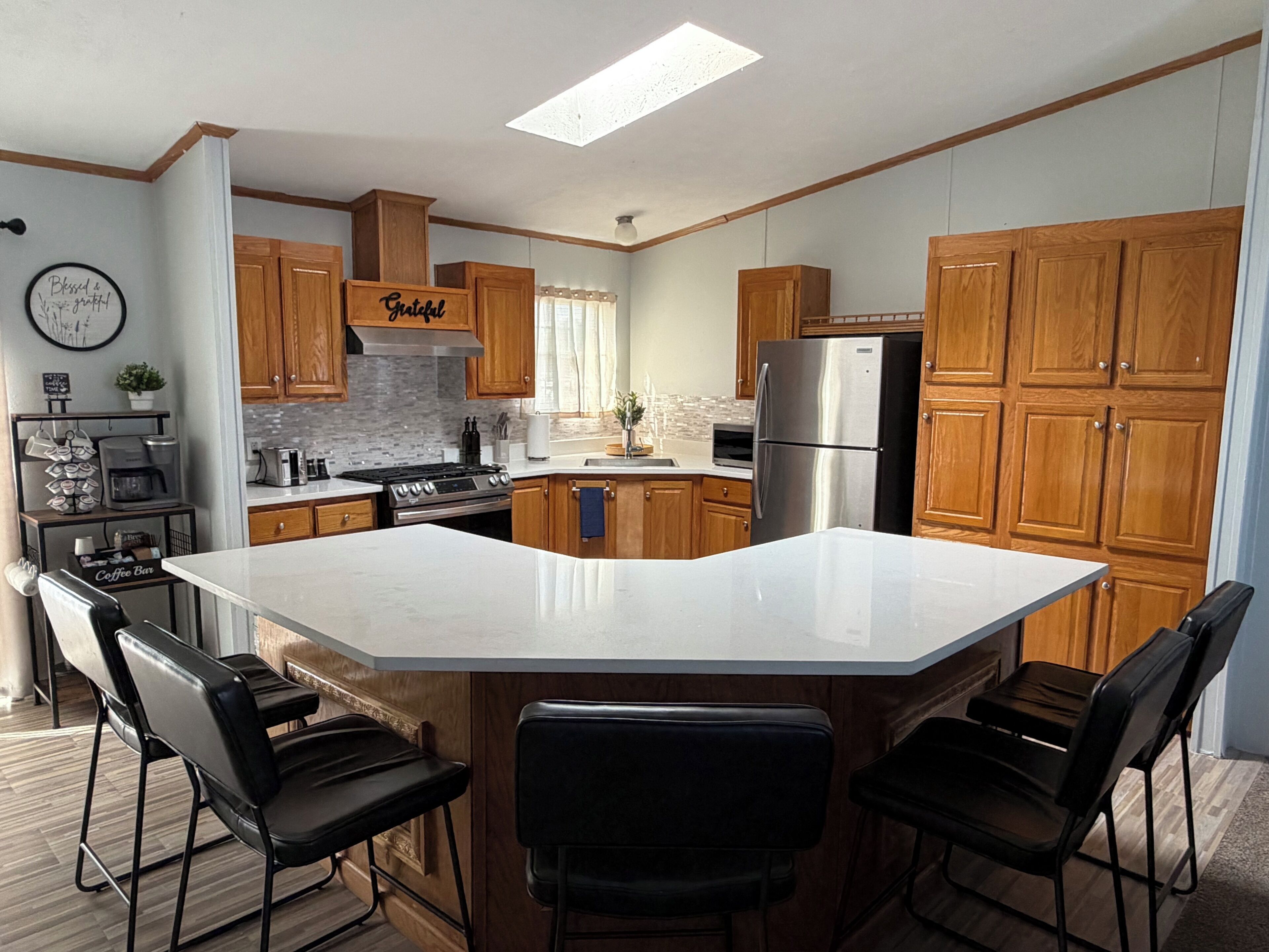 A modern kitchen with wooden cabinets, a stainless steel refrigerator, a central island with bar stools, and a coffee station.