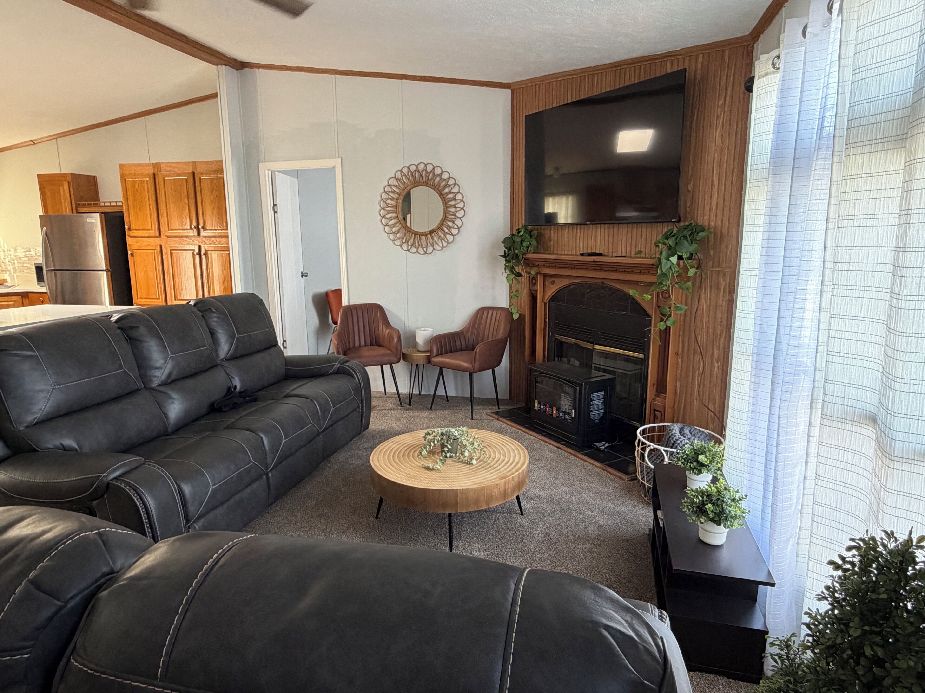 A living room with a black leather sofa, two brown chairs, a round coffee table, a flat-screen TV, a fireplace, and wooden cabinets.