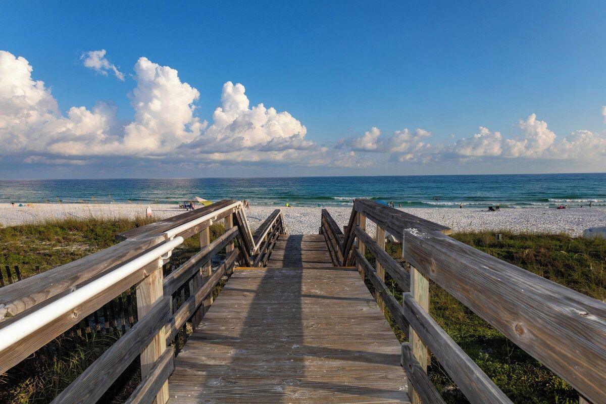 A wooden boardwalk leading to a beach with a clear sky and clouds.