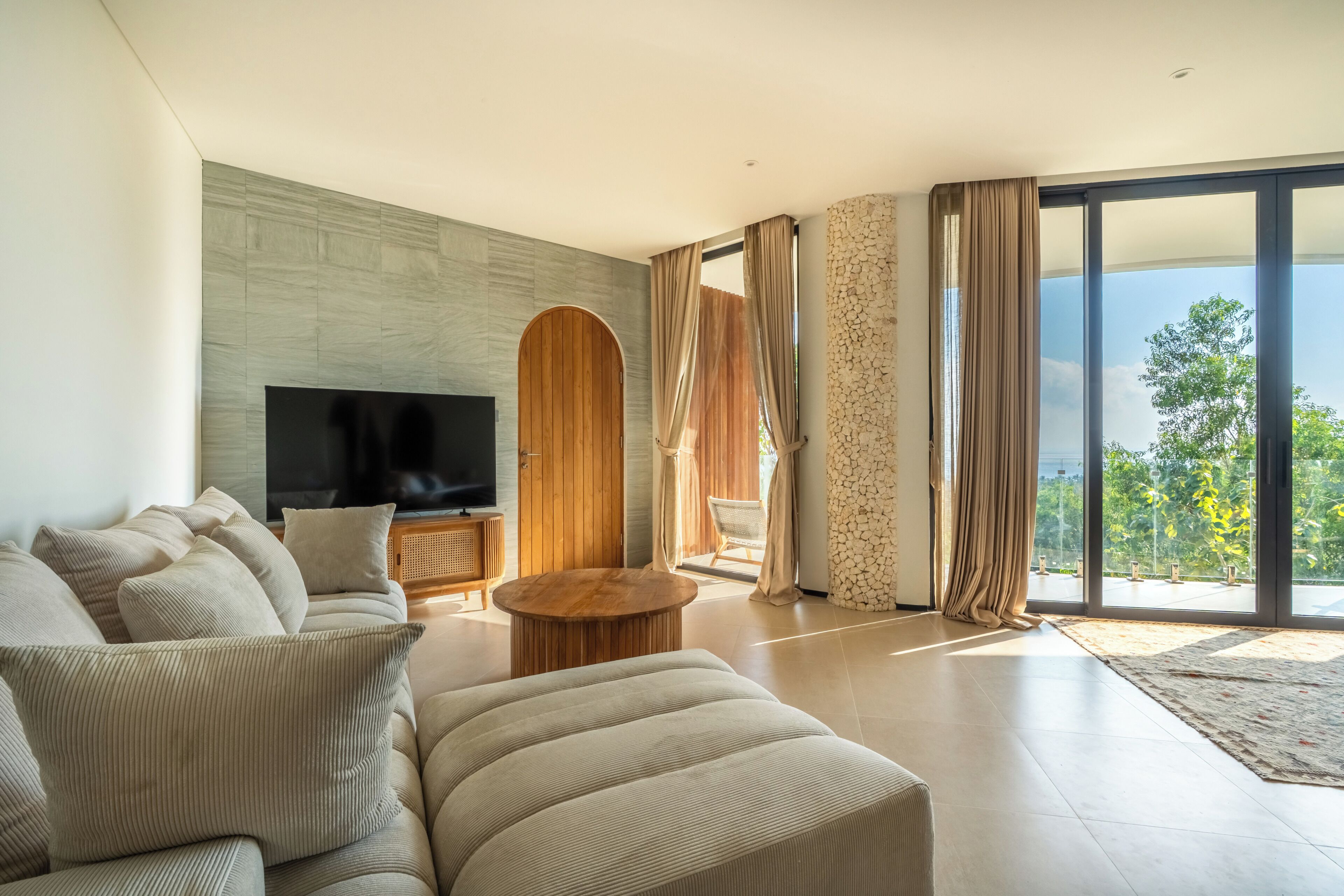 Una sala de estar moderna con un televisor de pantalla plana grande, un sofá, una mesa de centro y vistas a zonas verdes a través de amplios ventanales.