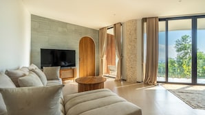 A modern living room with a large flat-screen TV, a sofa, a coffee table, and a view of greenery through large windows. - Villa Karilla (Penida Island)