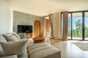 Una sala de estar moderna con un televisor de pantalla plana grande, un sofá, una mesa de centro y vistas a zonas verdes a través de amplios ventanales.