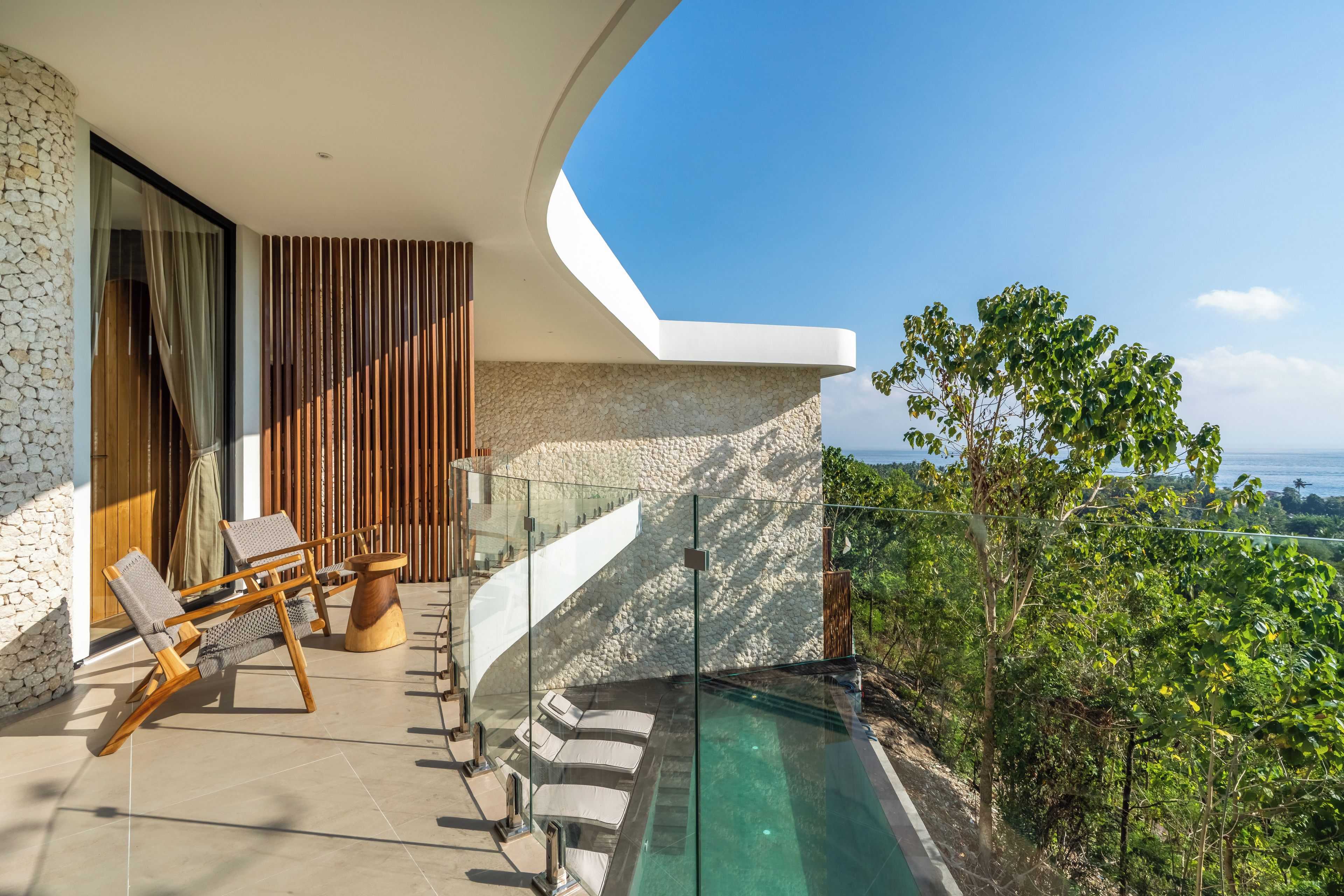 A modern outdoor balcony with a pool, wooden furniture, and a stone wall.