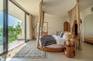 A modern bedroom with a large bed, a wooden bedside table, and a view of greenery outside.