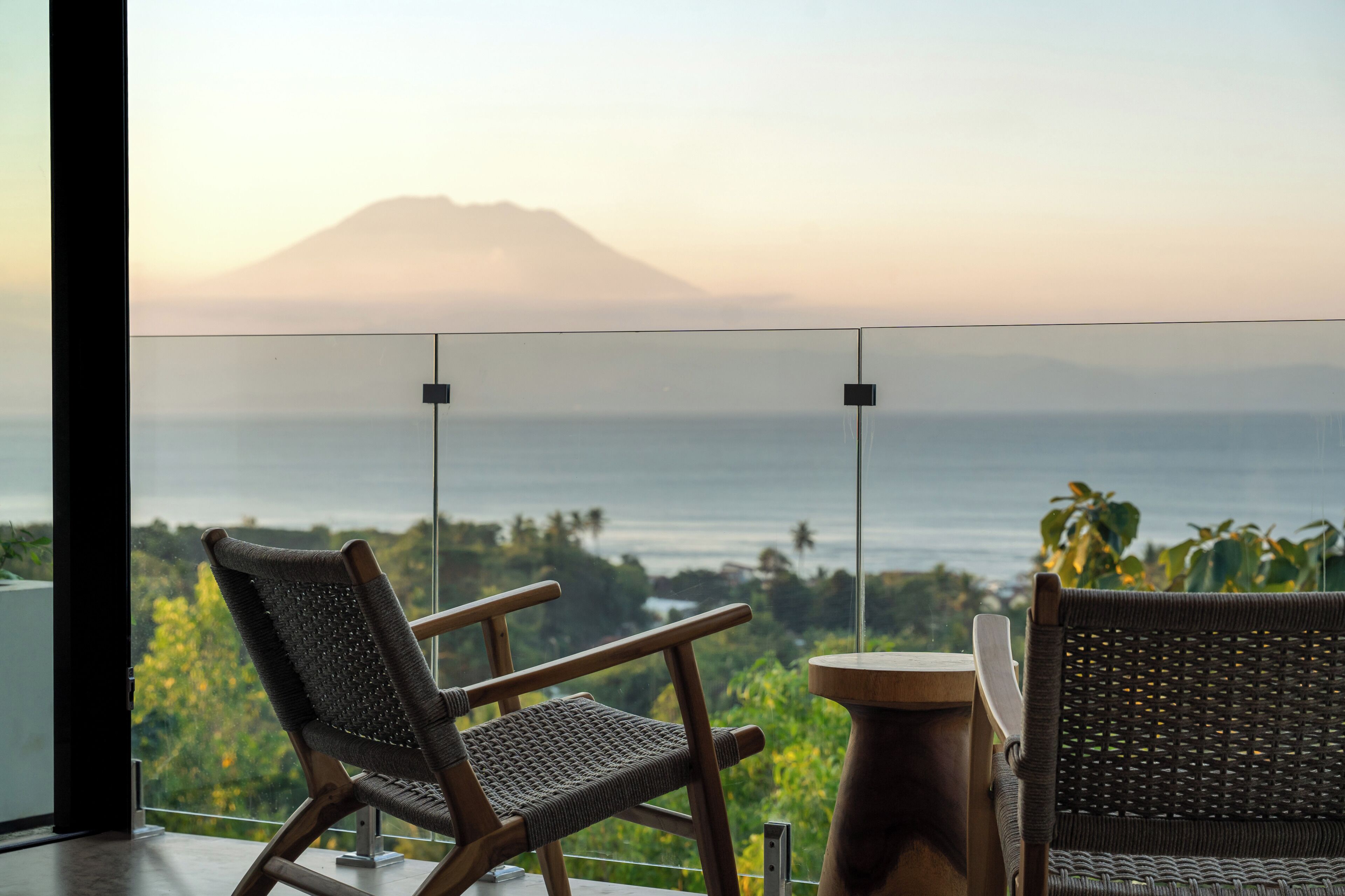 A room with a view of a mountain and the sea, featuring two wicker chairs and a small wooden table.