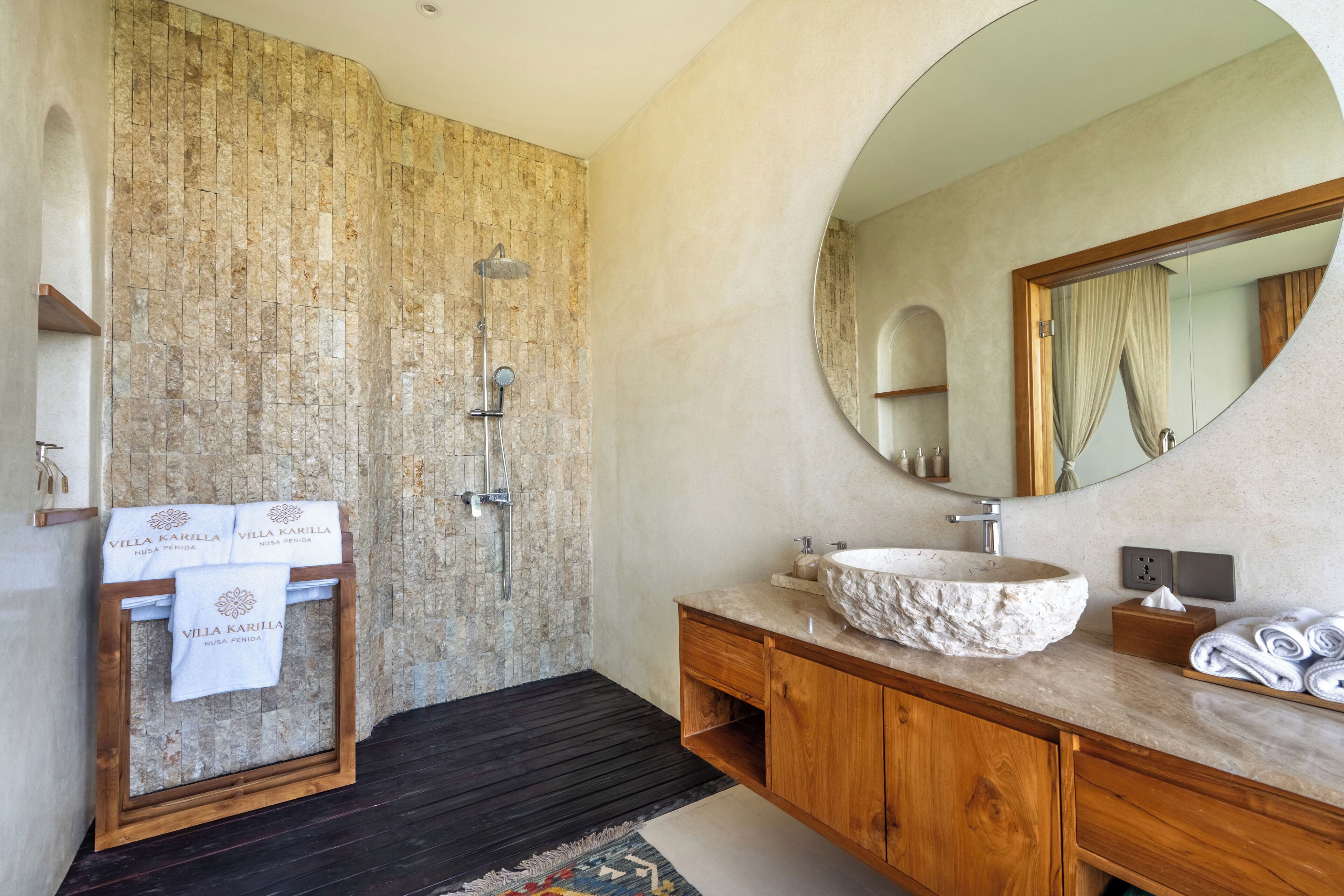 Une salle de bain avec un mur en pierre, un meuble-lavabo en bois, un miroir rond et une baignoire sur pieds.