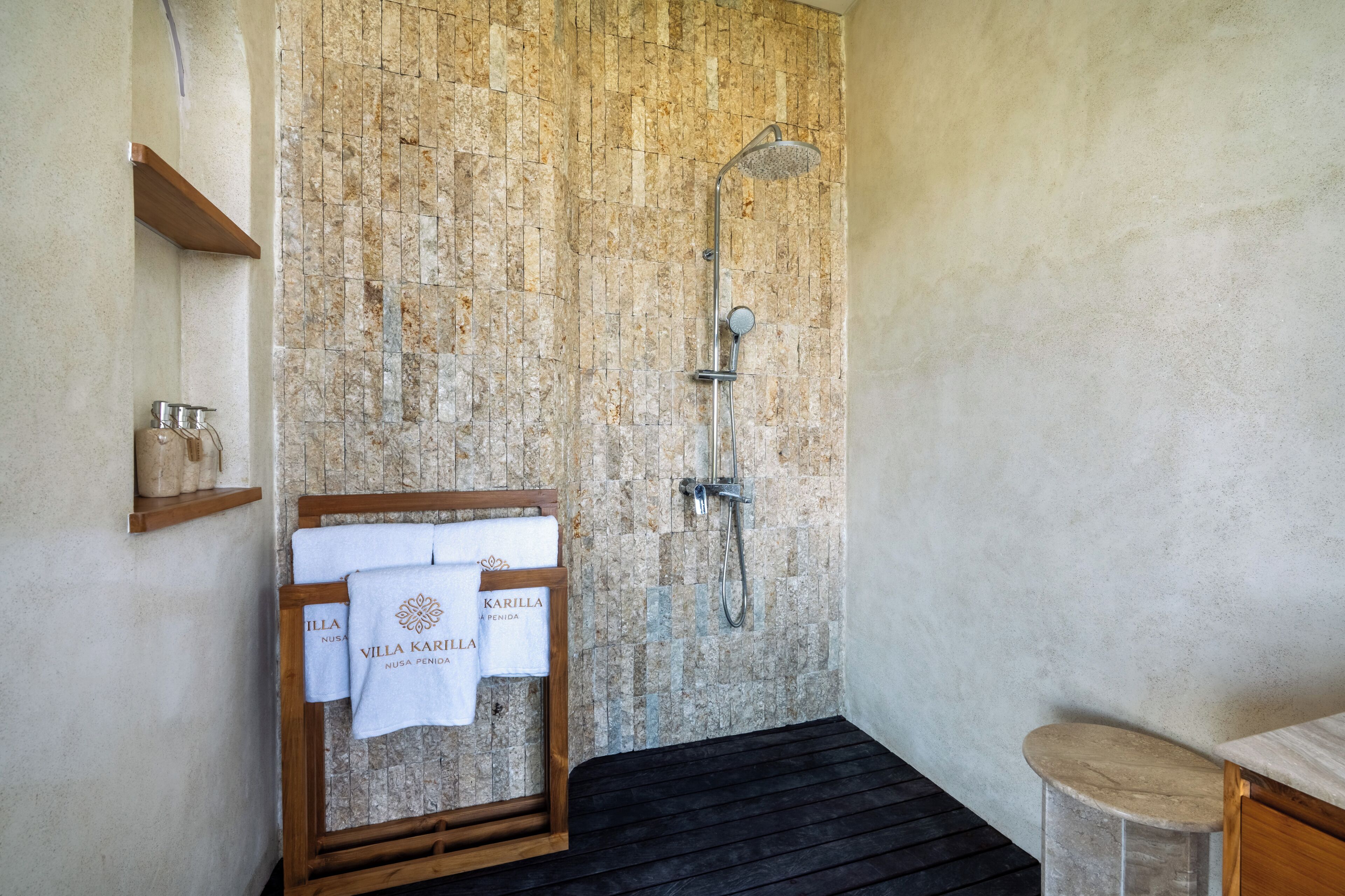 Un baño con pared de piedra, ducha, banco de madera con toallas y un estante peque ño con recipientes.