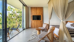 A modern bedroom with a large bed, a TV mounted on a wooden panel, a balcony with wicker furniture, and a view of greenery. - Villa Karilla (Penida Island)