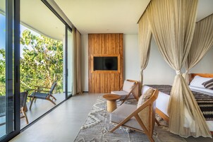 A modern bedroom with a large bed, a TV mounted on a wooden panel, a balcony with wicker furniture, and a view of greenery.