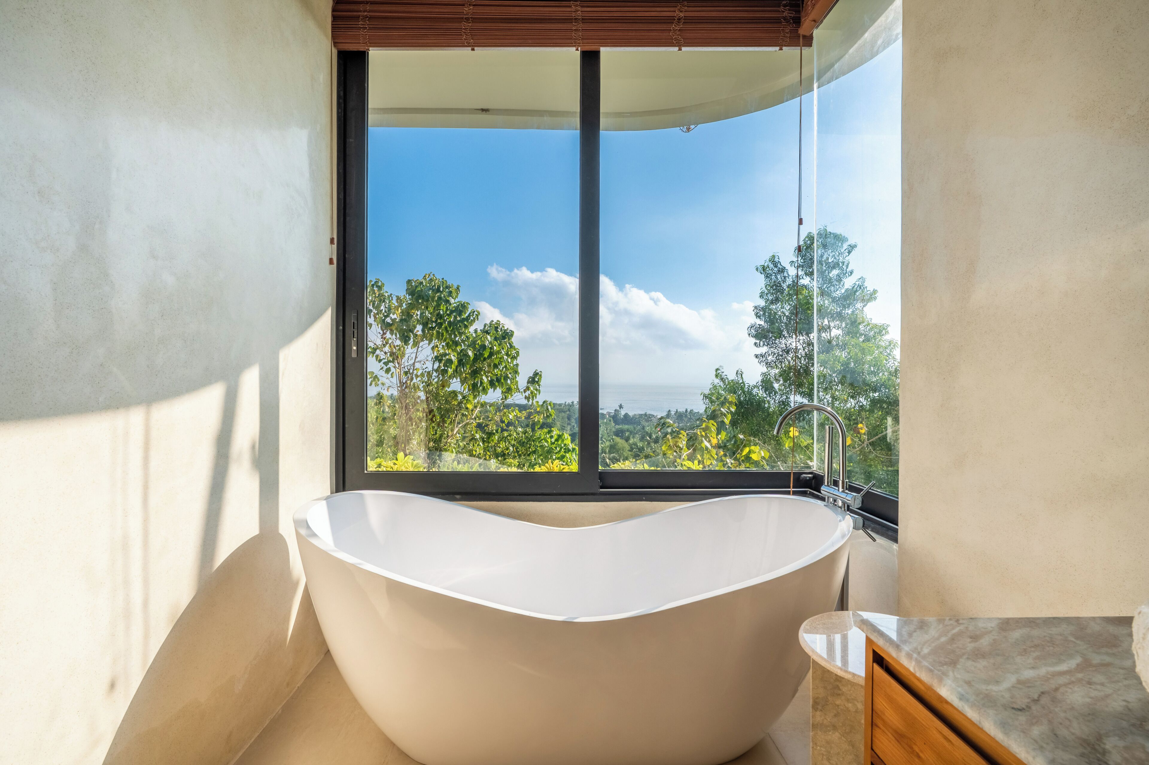 A modern bathroom with a freestanding bathtub, a window with a view of trees and a clear sky, and a wooden vanity.